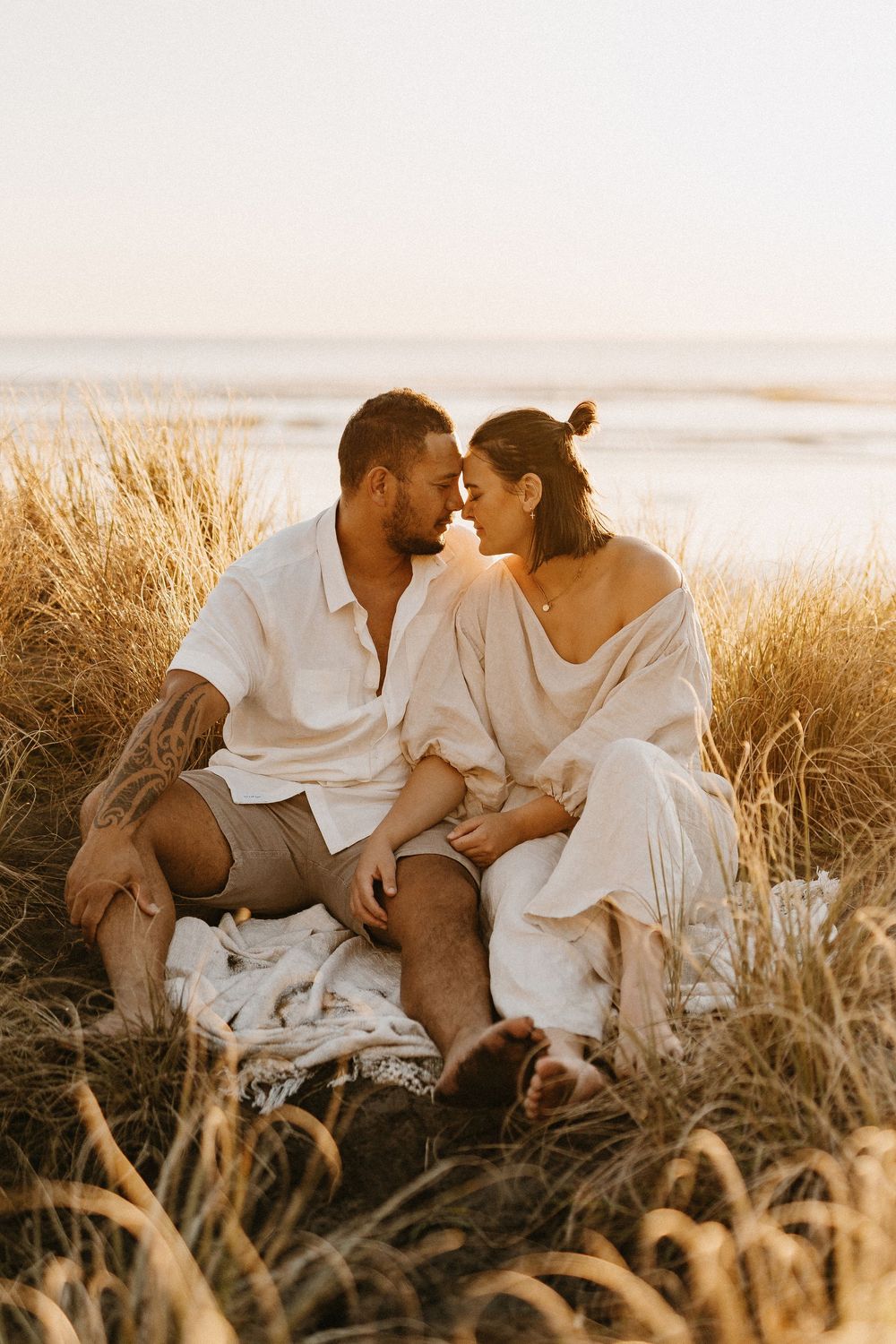 A romantic couple in white clothing sits together on a blanket among tall grass during golden hour at the beach.