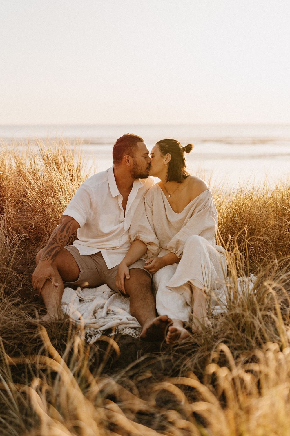 Couple sitting together on a beach at sunset surrounded by golden wheat grass.