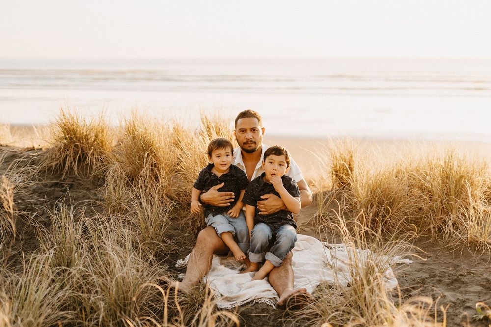 Family sits together on a blanket among tall golden grasses along the beach at sunset.