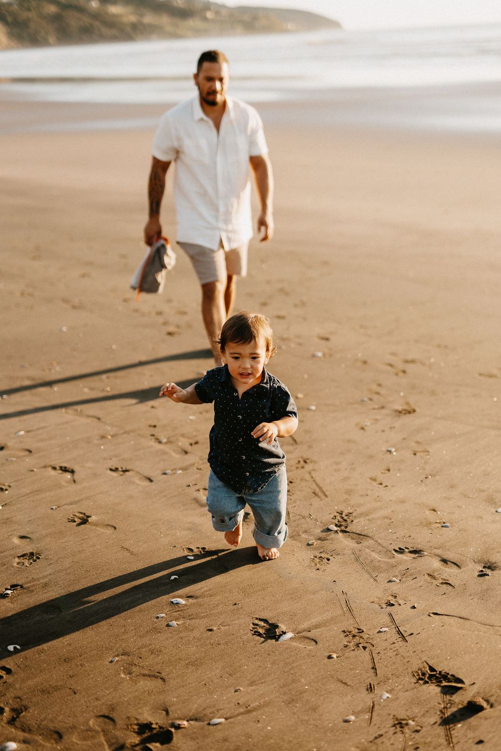 A joyful child plays with their parent on a sunlit sandy beach while footprints mark their path along the shore.
