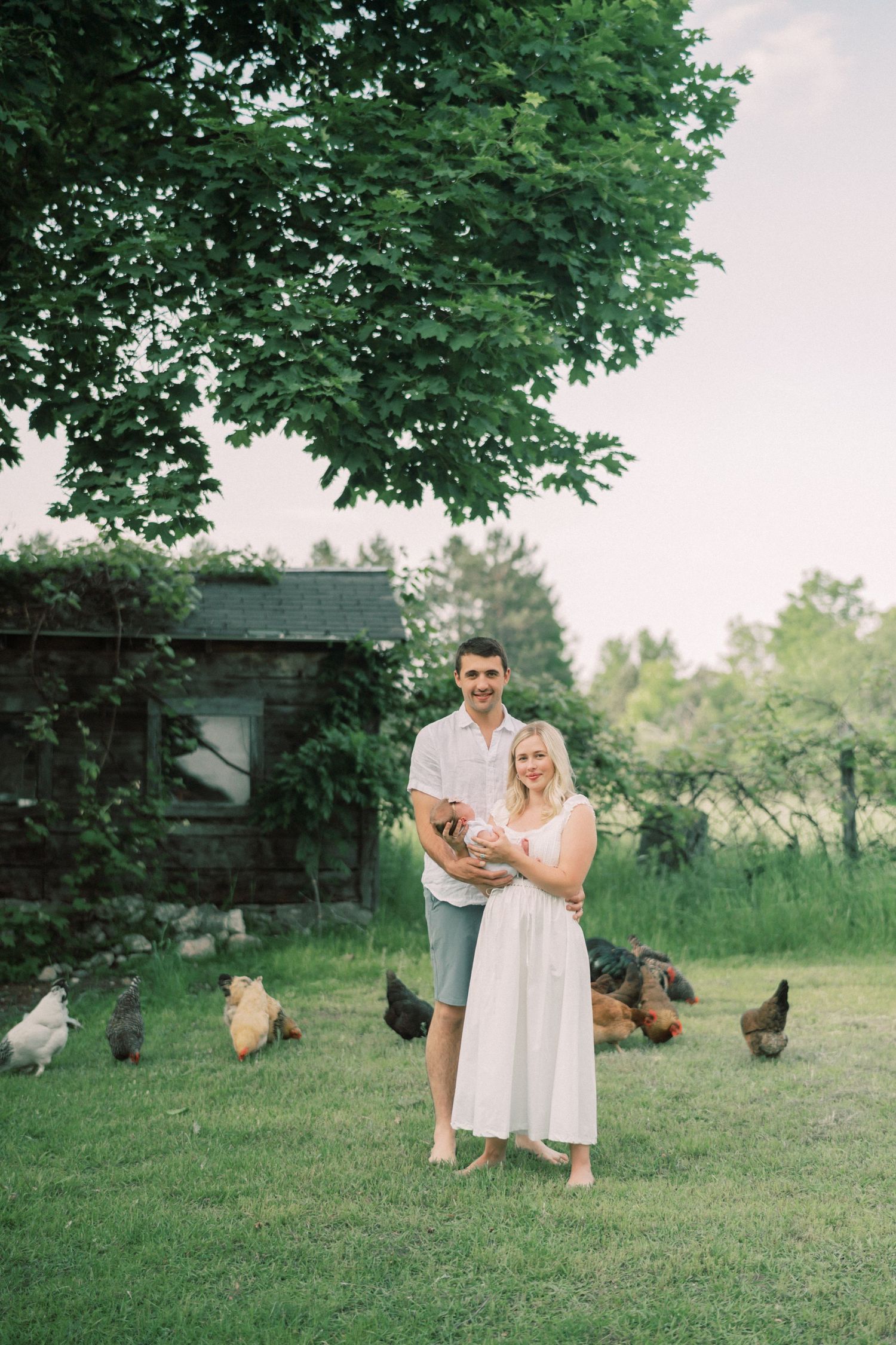 A couple stands together on a green lawn with chickens roaming around them near a rustic outbuilding on a cloudy day.