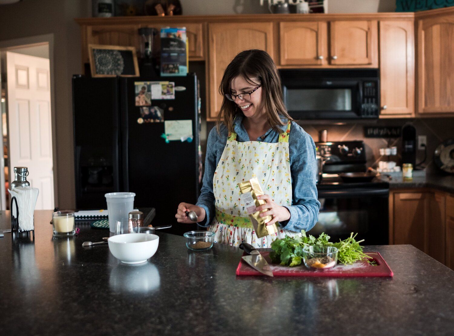 A person in glasses and denim jacket preparing fresh salad ingredients at a dark countertop in a home kitchen.