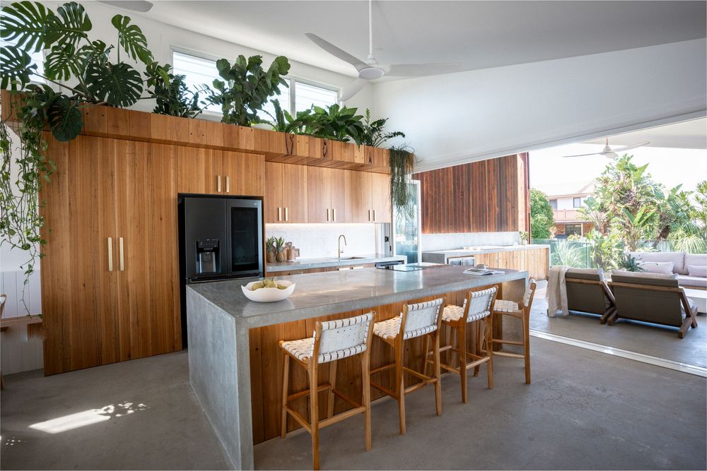 Modern open kitchen with warm wood cabinets, concrete floors, marble island and greenery on top of cabinets.