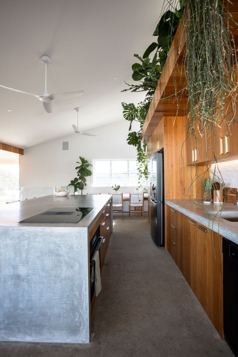 Modern kitchen with concrete floors, wooden cabinets, hanging plants and white ceiling fans in a minimalist design.