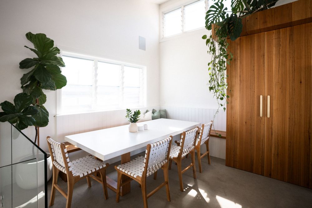 Modern dining room with white table, woven chairs, wooden storage cabinet and potted plants near bright windows.