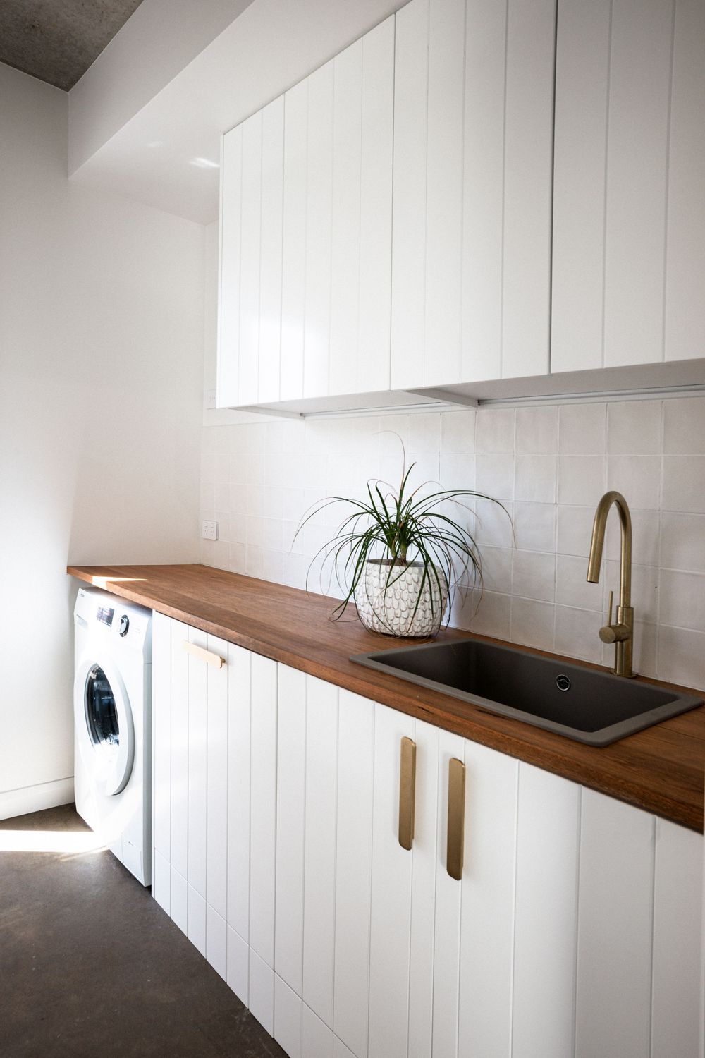 Modern white laundry room with wooden countertop, white cabinets, and gold hardware features an inset washer and dryer.