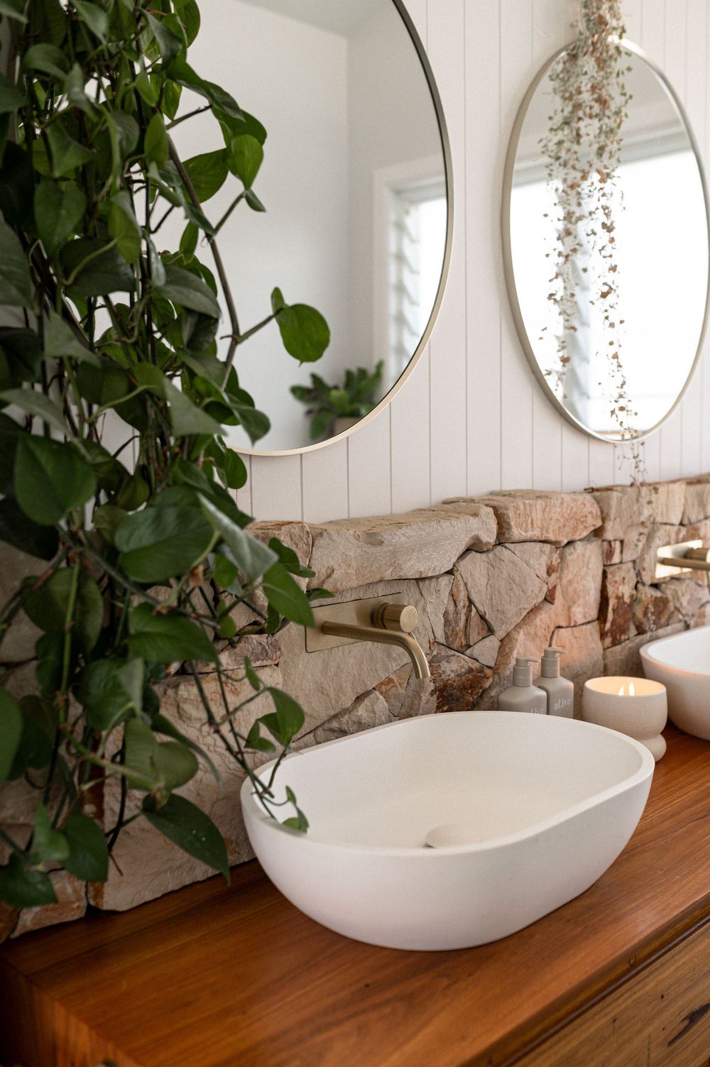 Modern bathroom vanity with white vessel sink on wooden counter featuring natural stone wall and oval mirror.