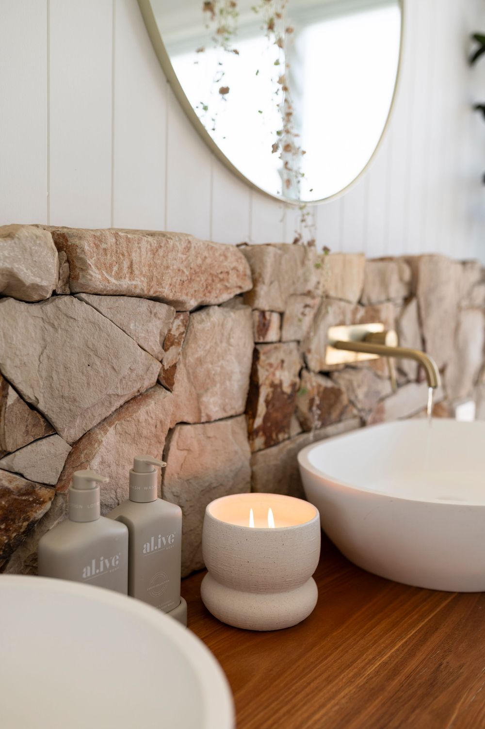 Modern bathroom vanity with stone wall backsplash, round mirror, white vessel sinks and lit candle on wooden counter.