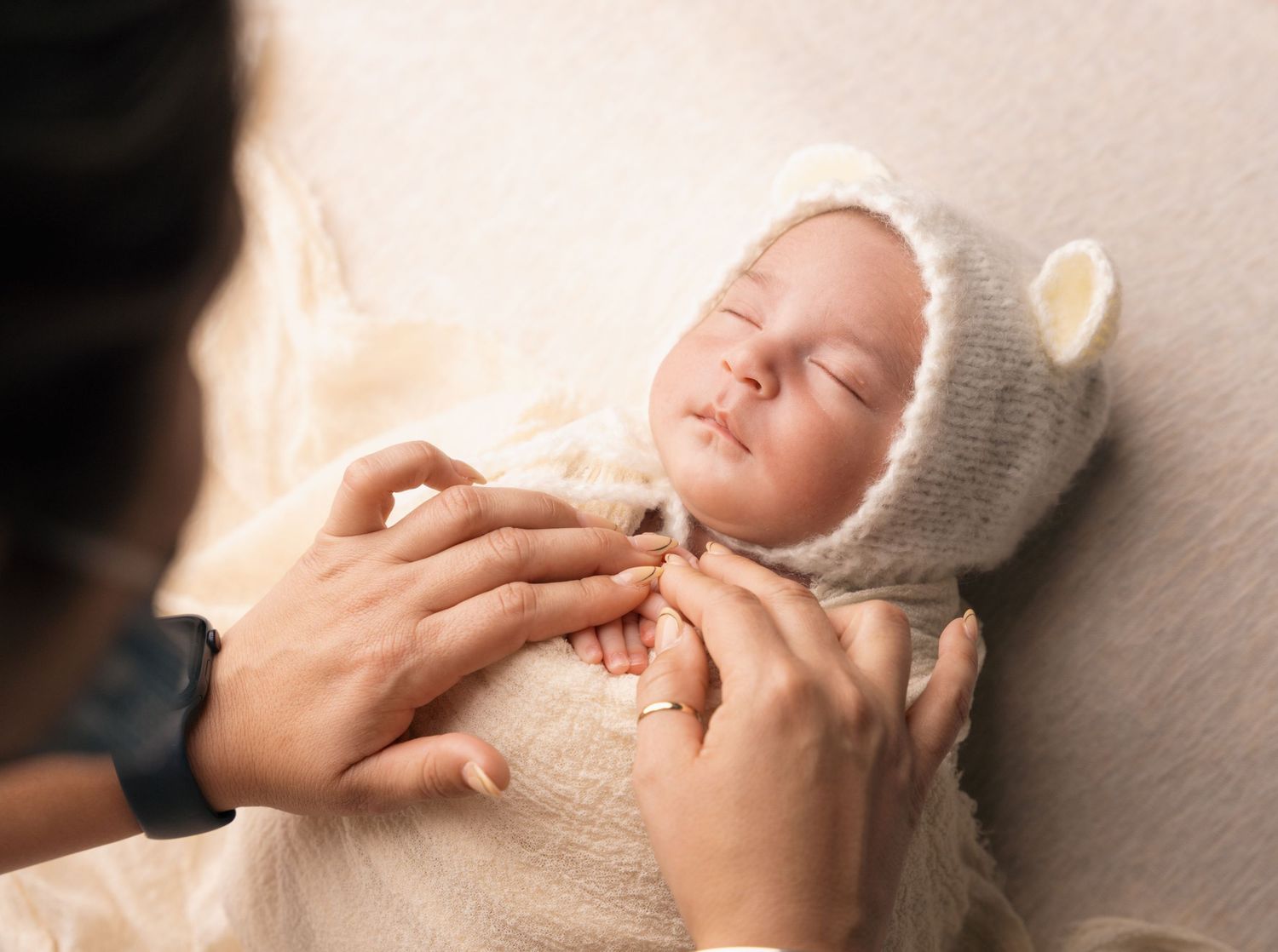 A sleeping baby wearing a knit bonnet lies peacefully while gentle hands adjust their soft white swaddle.