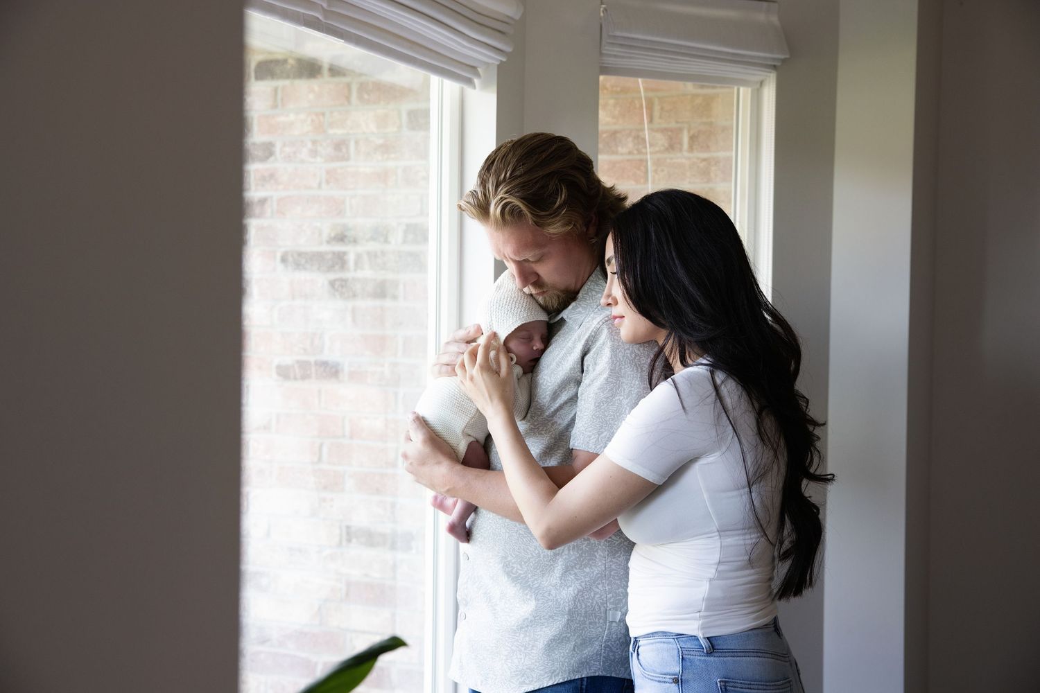 Couple embrace with newborn baby girl near a window in soft natural light in their home