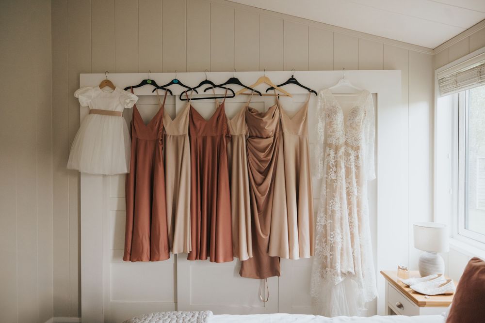 A collection of elegant bridesmaid dresses in neutral tones hung on hangers against a beige wall.