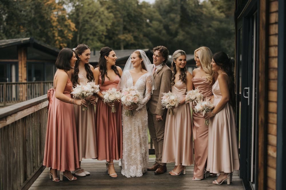 A wedding party in dusty rose and neutral colored dresses poses together on a wooden deck surrounded by trees.