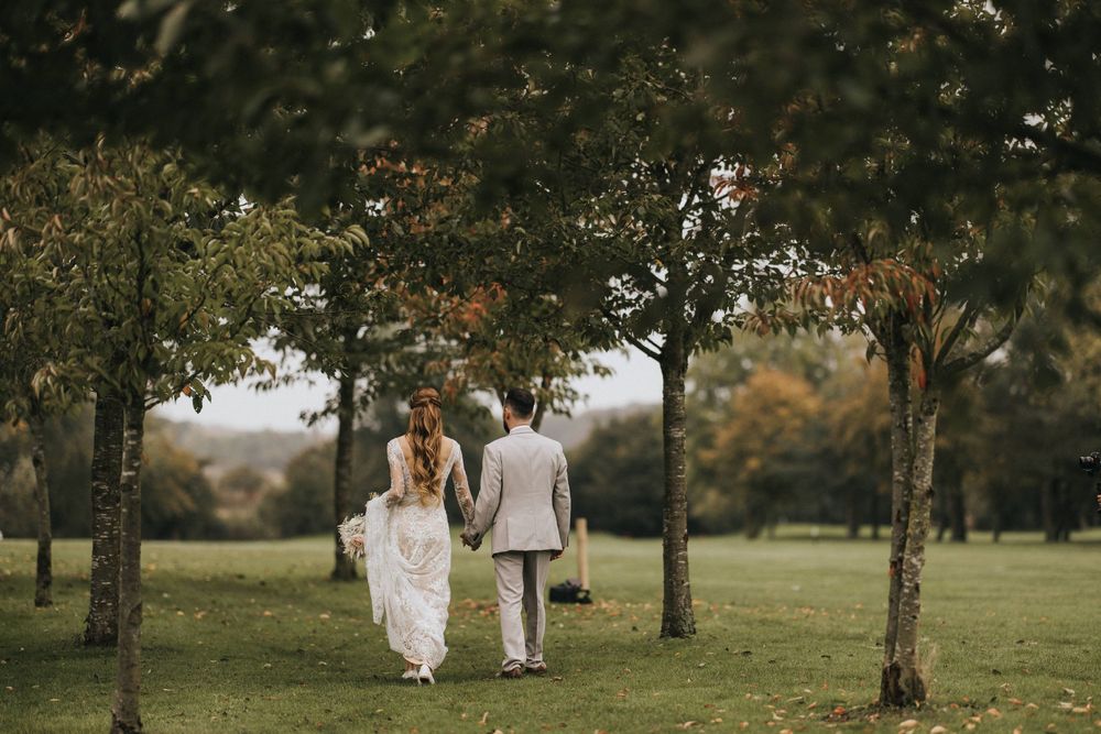 Bride and groom walk hand in hand through an autumn orchard with golden sunlight peeking through the trees.