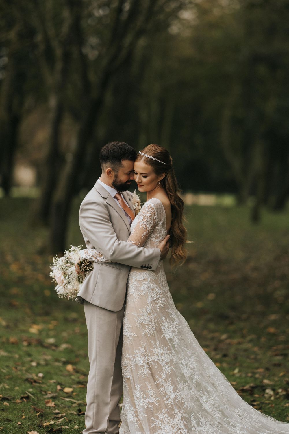 A romantic wedding couple embraces in a wooded setting with fall foliage and a beautiful lace dress with white bouquet.