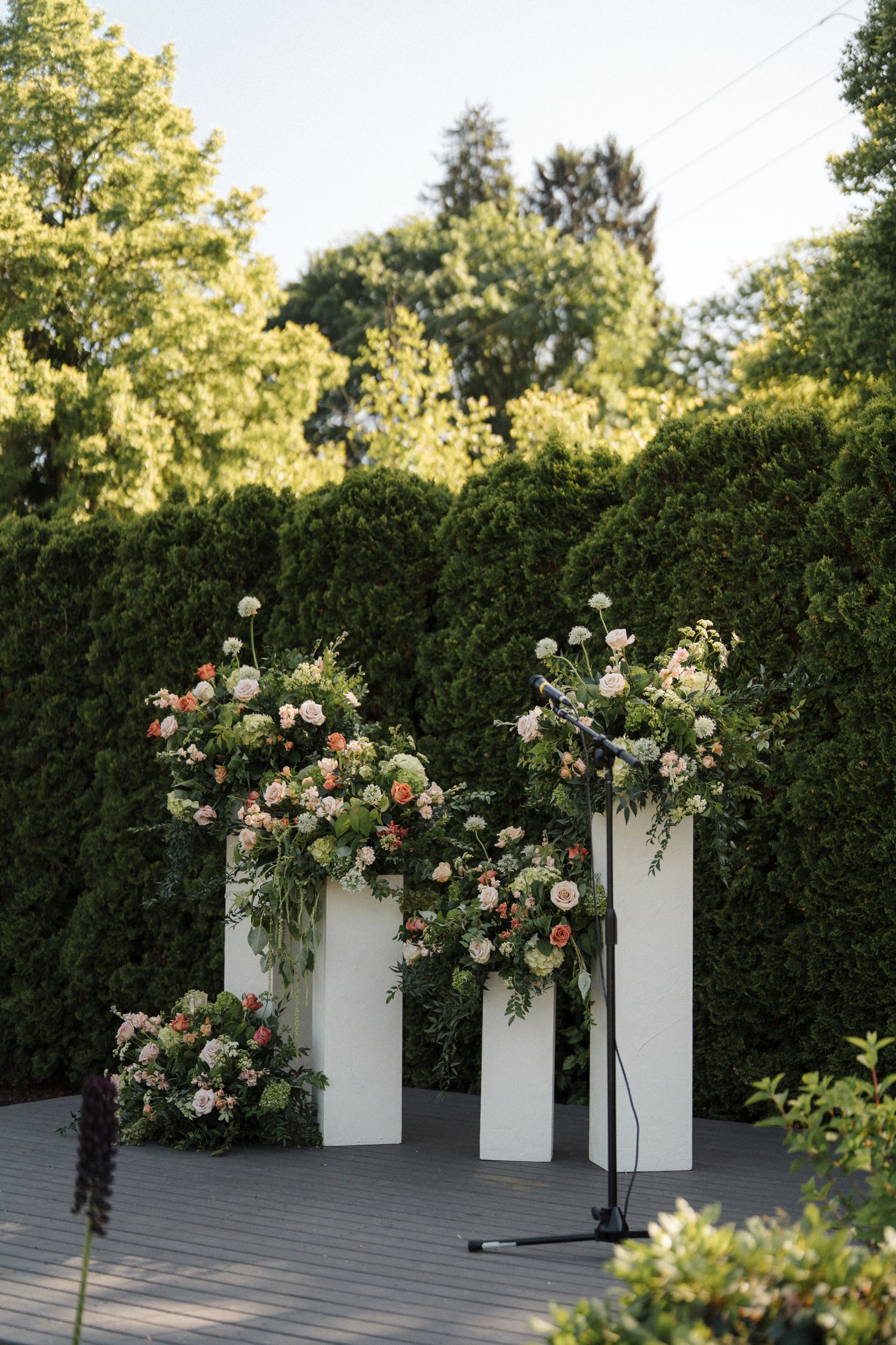 Three white pillars decorated with pink and peach floral arrangements stand against a green hedge backdrop in an outdoor setting.