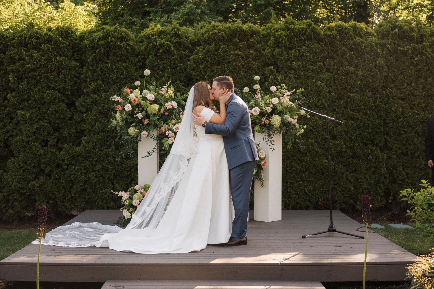A romantic outdoor wedding ceremony takes place against a hedge wall backdrop with white floral arrangements and pillars.