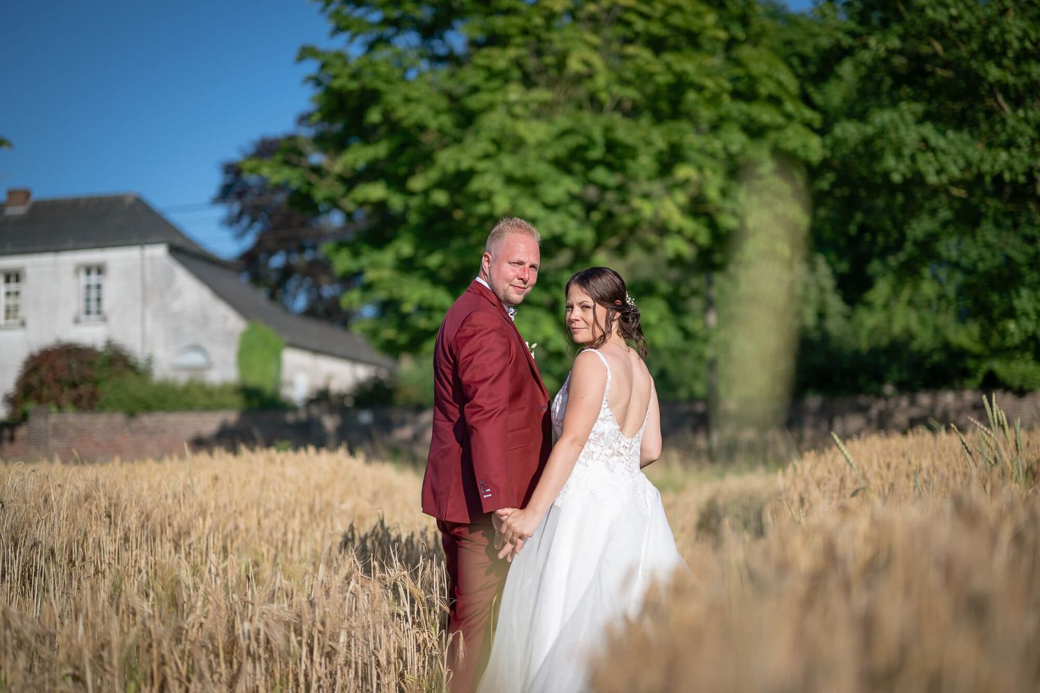 A couple in wedding attire stands intimately in a golden wheat field on a sunny summer day.
