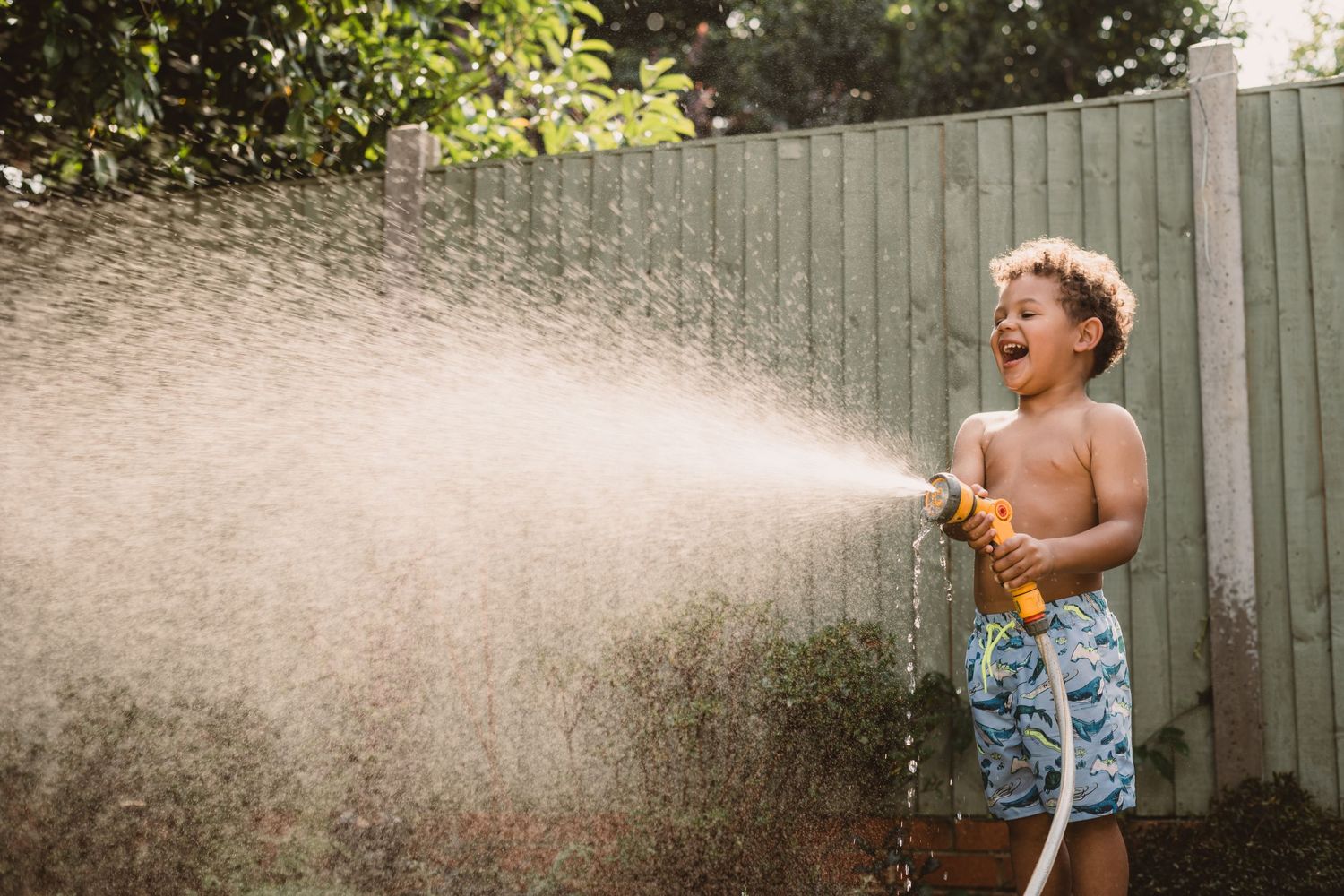 A joyful young child plays with a water hose on a hot summer day, spraying water in the backyard.