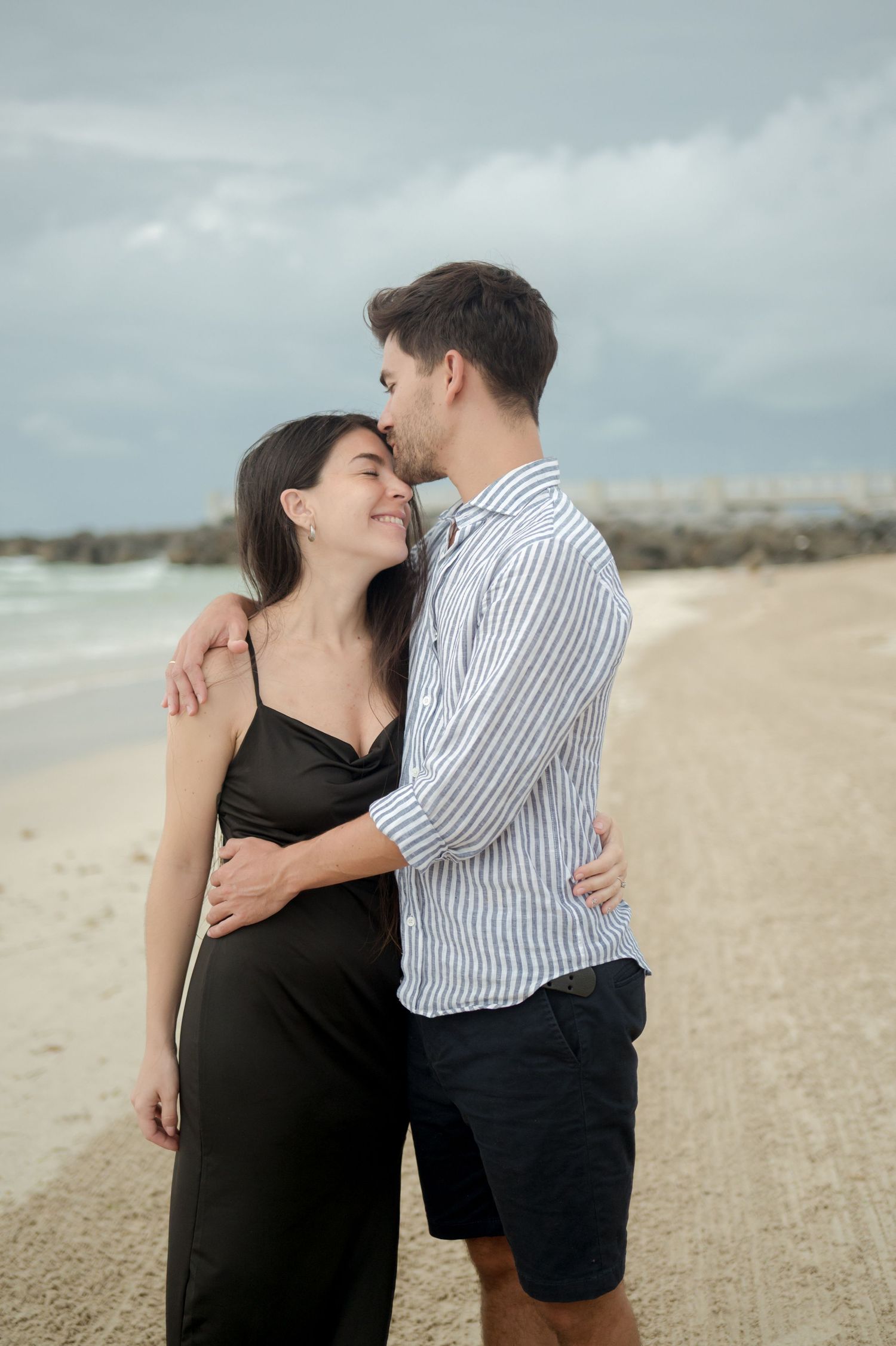 A romantic couple embraces on a cloudy beach, wearing a black dress and striped shirt respectively.