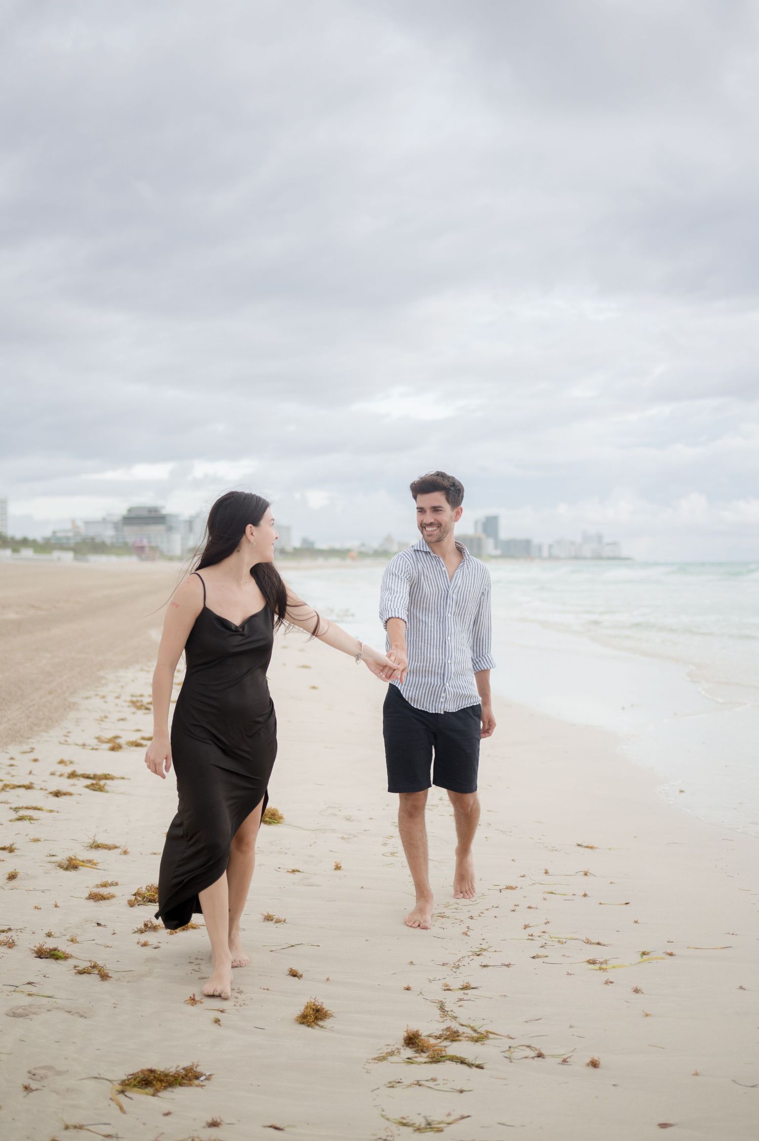 A couple walks together barefoot on a cloudy beach, with the woman wearing a black dress and the man in casual attire.