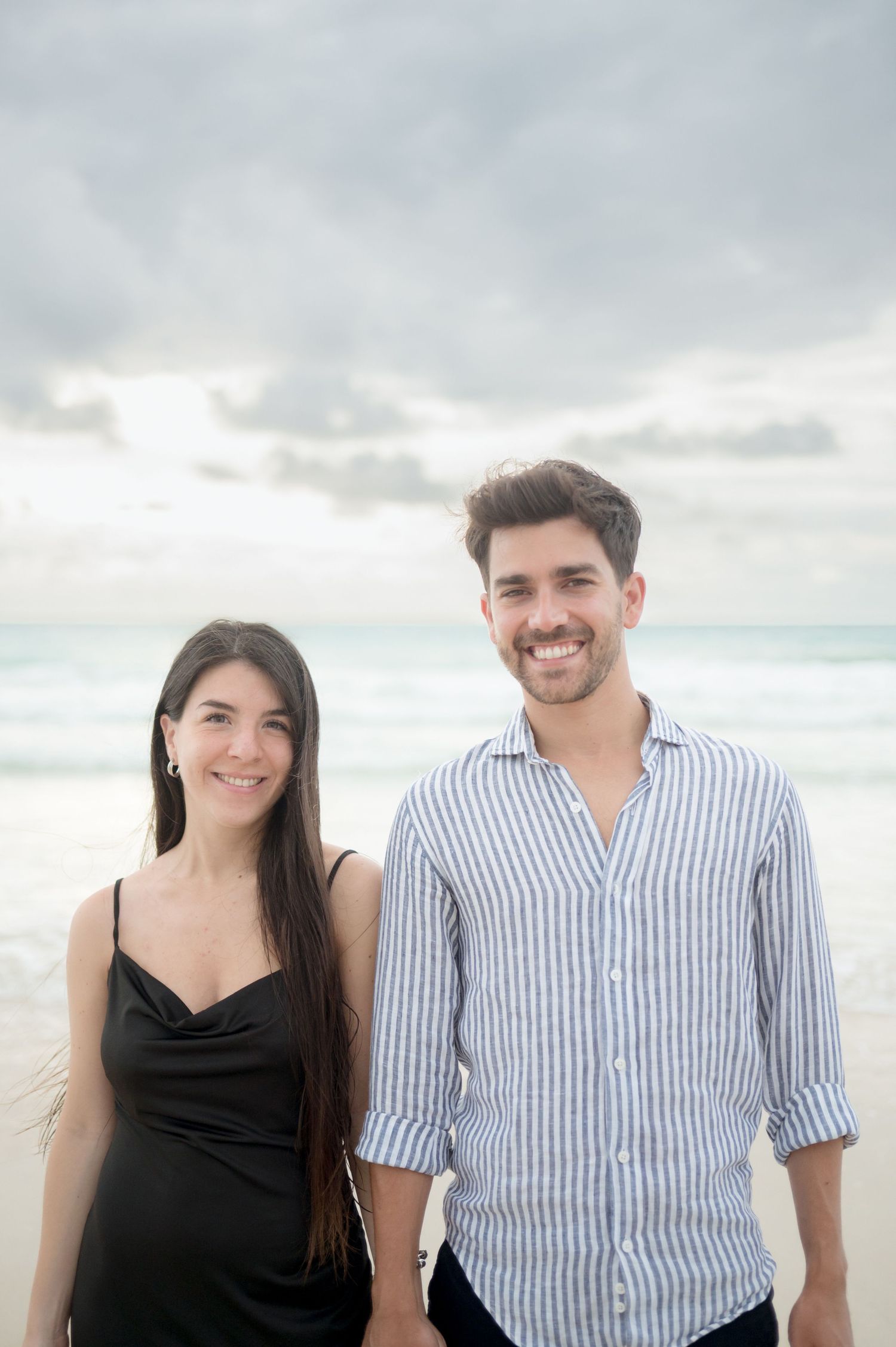 Couple in coordinated black dress and blue striped shirt outfits standing together on a beach with cloudy sky.