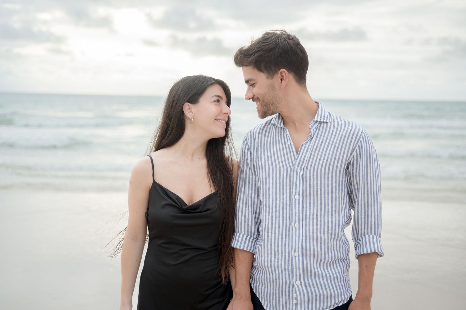 A couple in black dress and striped shirt enjoying a romantic moment together on a cloudy beach.