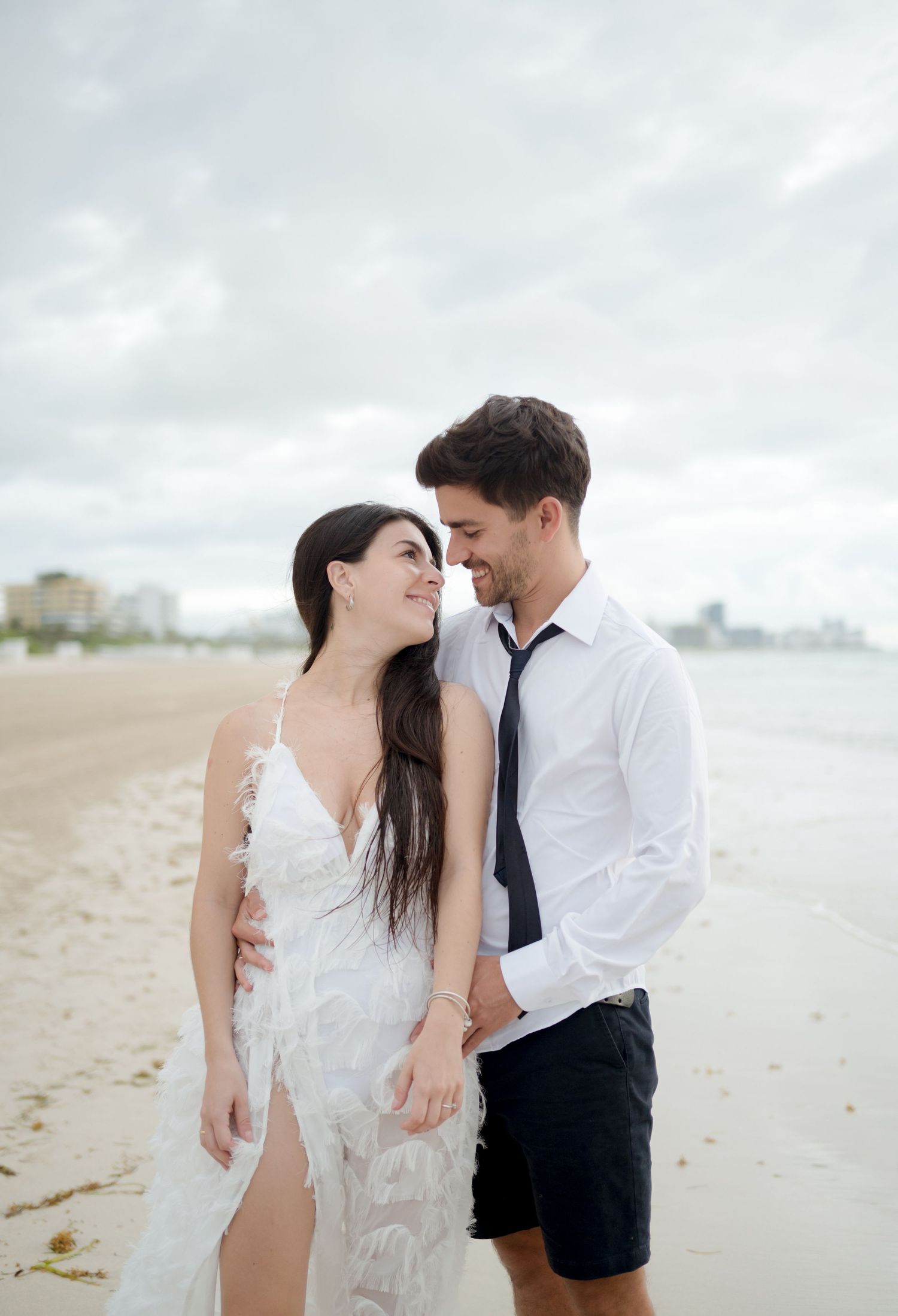 A romantic couple in formal attire share an intimate moment while walking along a misty beach shore.