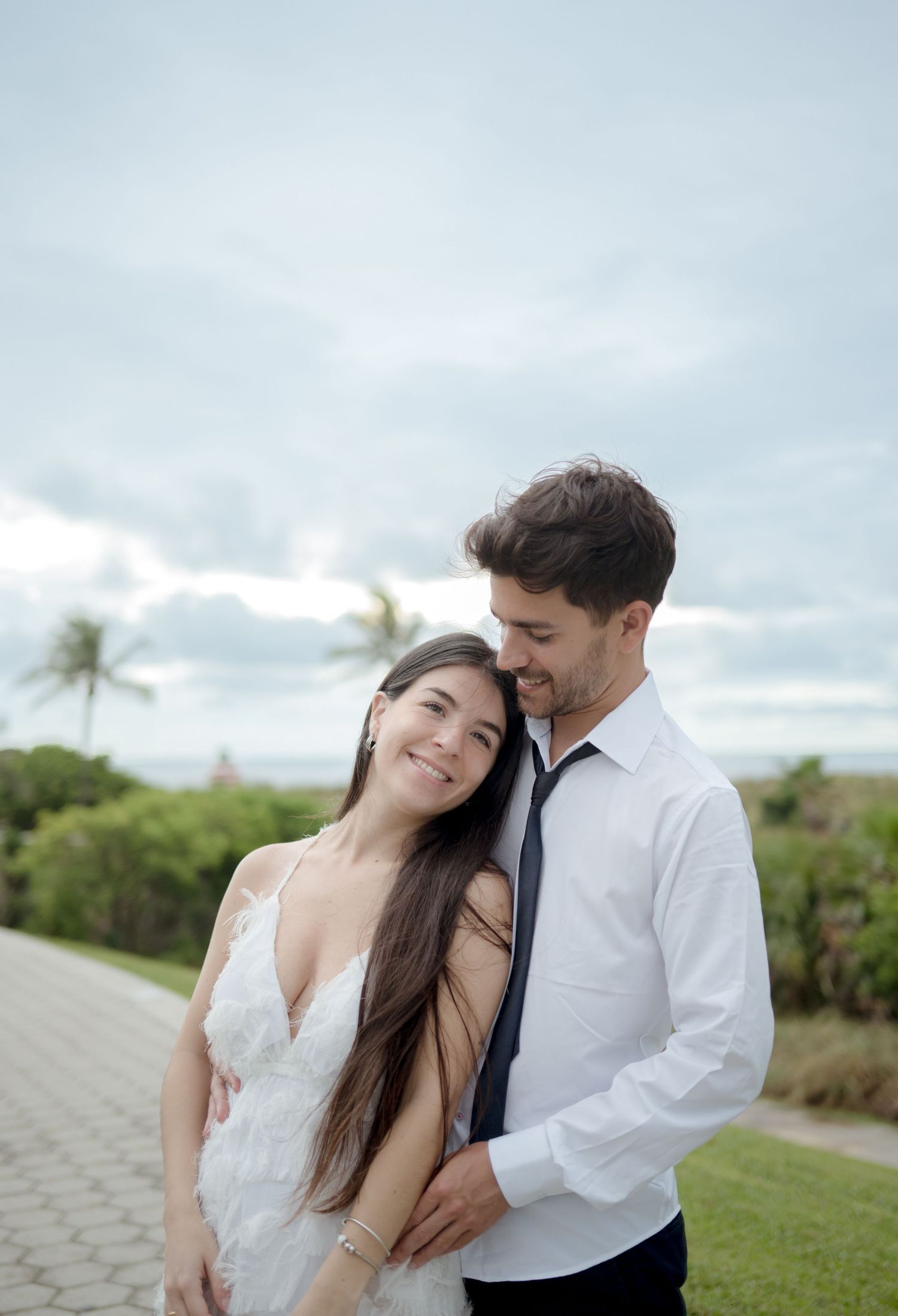 A romantic couple embraces outdoors against a cloudy sky with palm trees and greenery in the background.