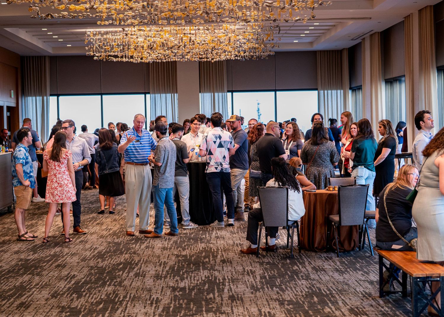 Large group mingling in an elegant event space with crystal chandelier and panoramic windows.