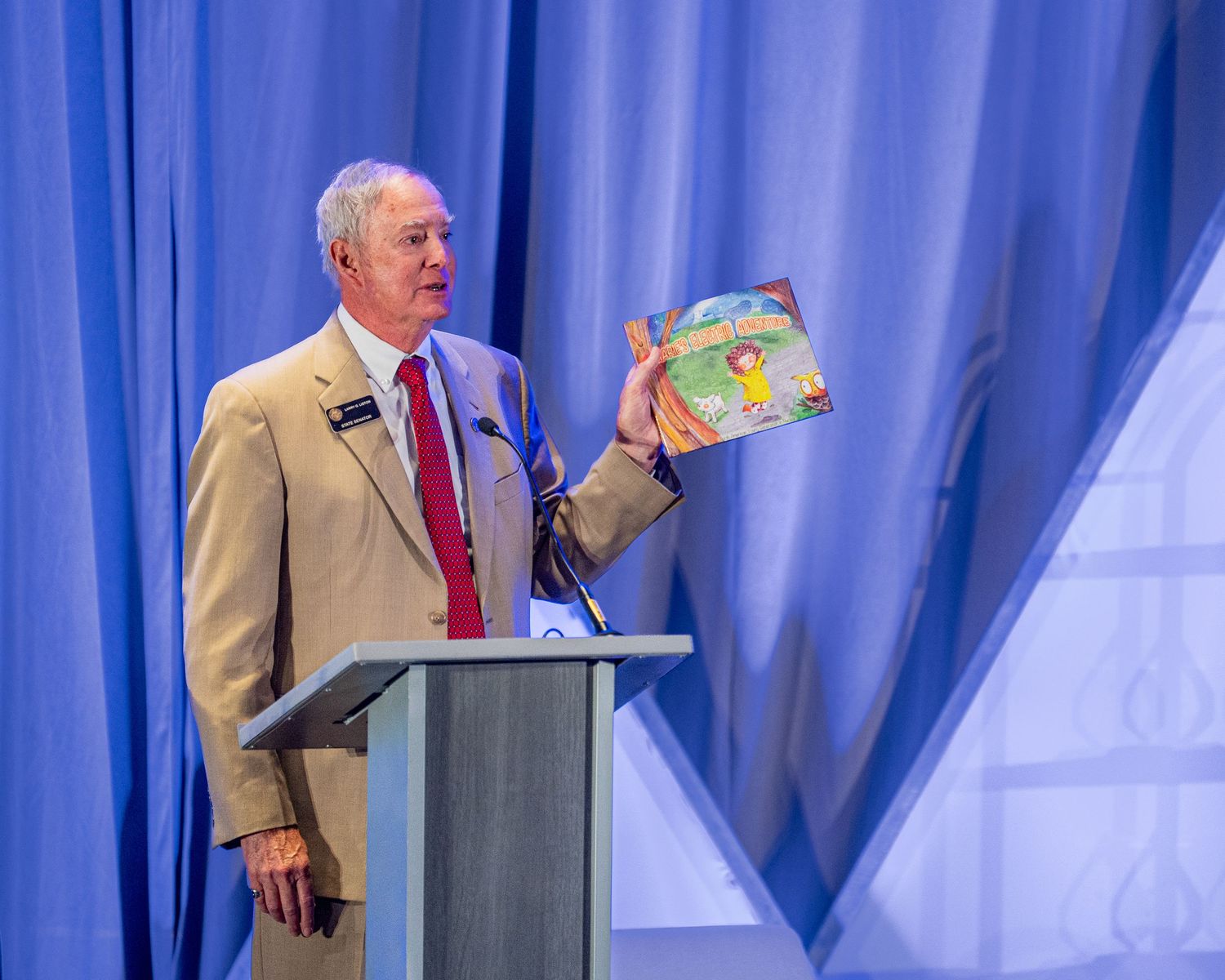 Speaker presenting at a podium with blue stage lighting and curtains.