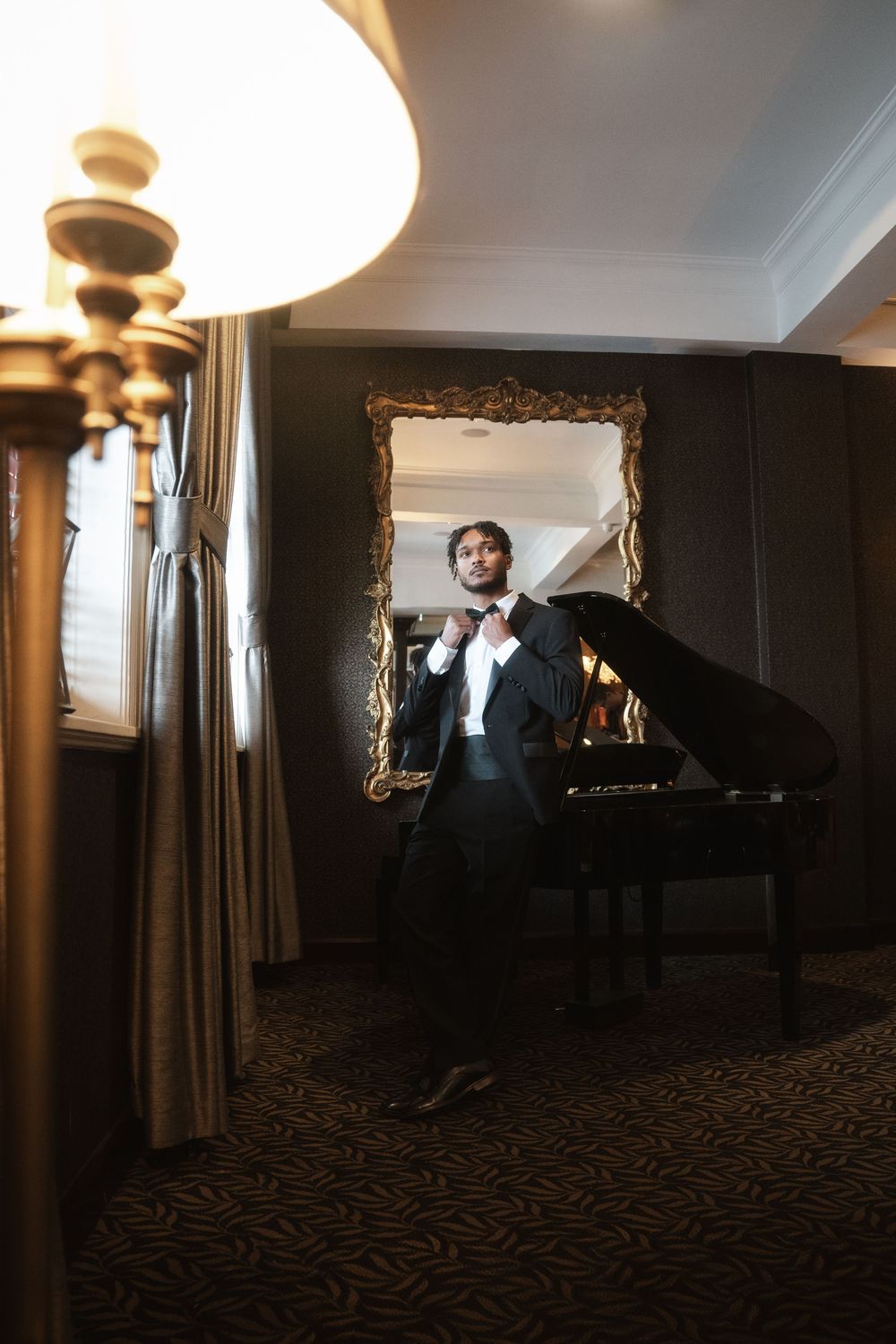 An elegant musician in formal attire adjusts their bowtie while standing next to a grand piano in a dimly lit room.