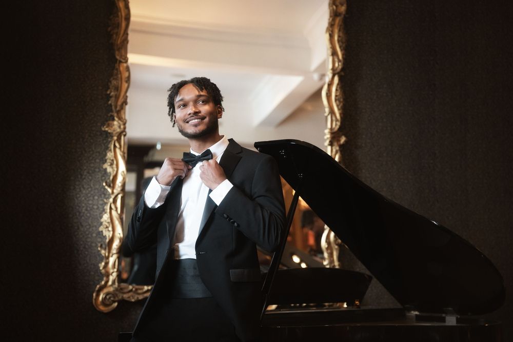 A smiling performer in a black tuxedo and bowtie adjusts their tie while standing by a grand piano.