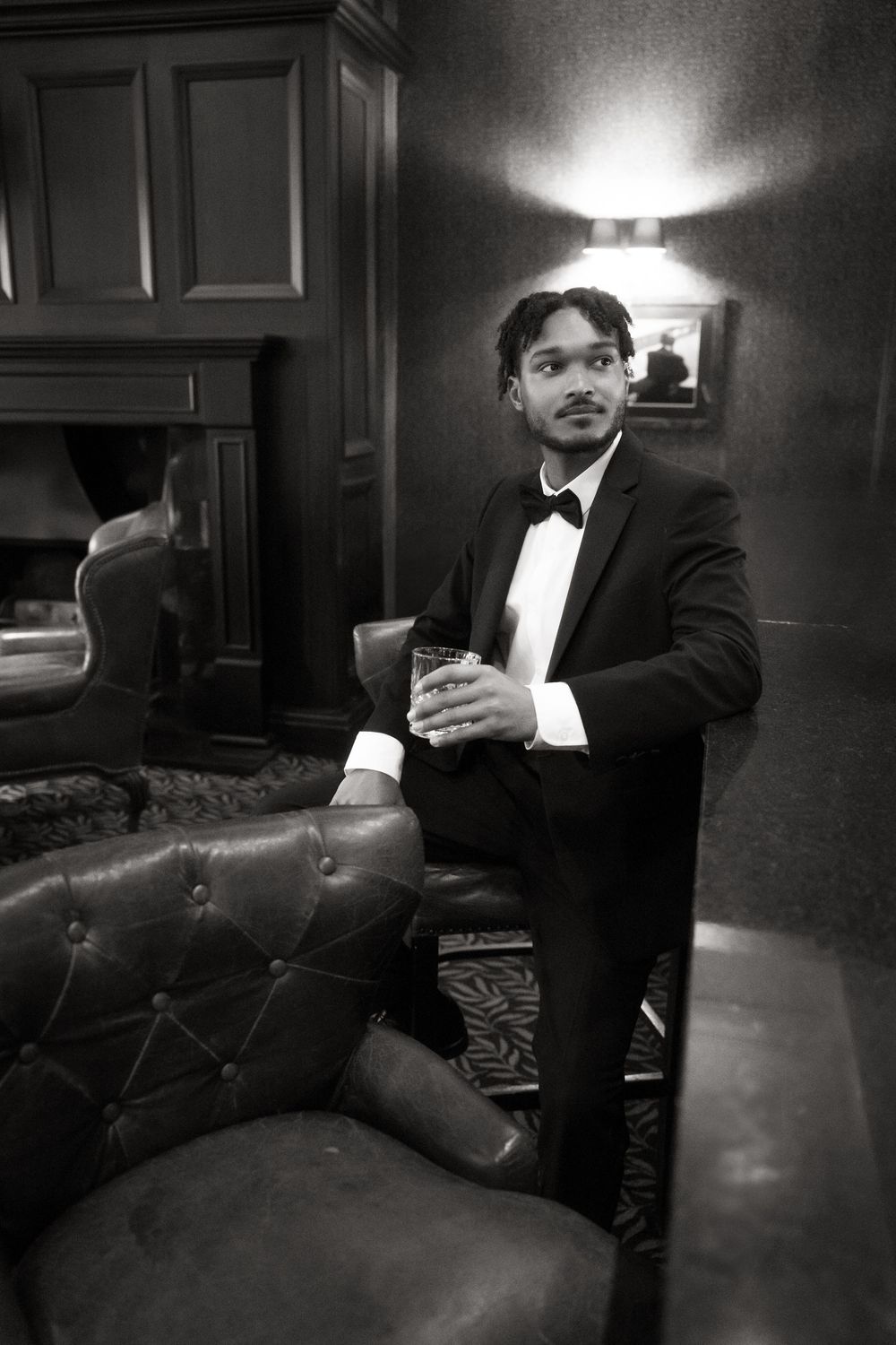 A person in formal black tie attire sits casually on a leather chair in a dimly lit elegant room.