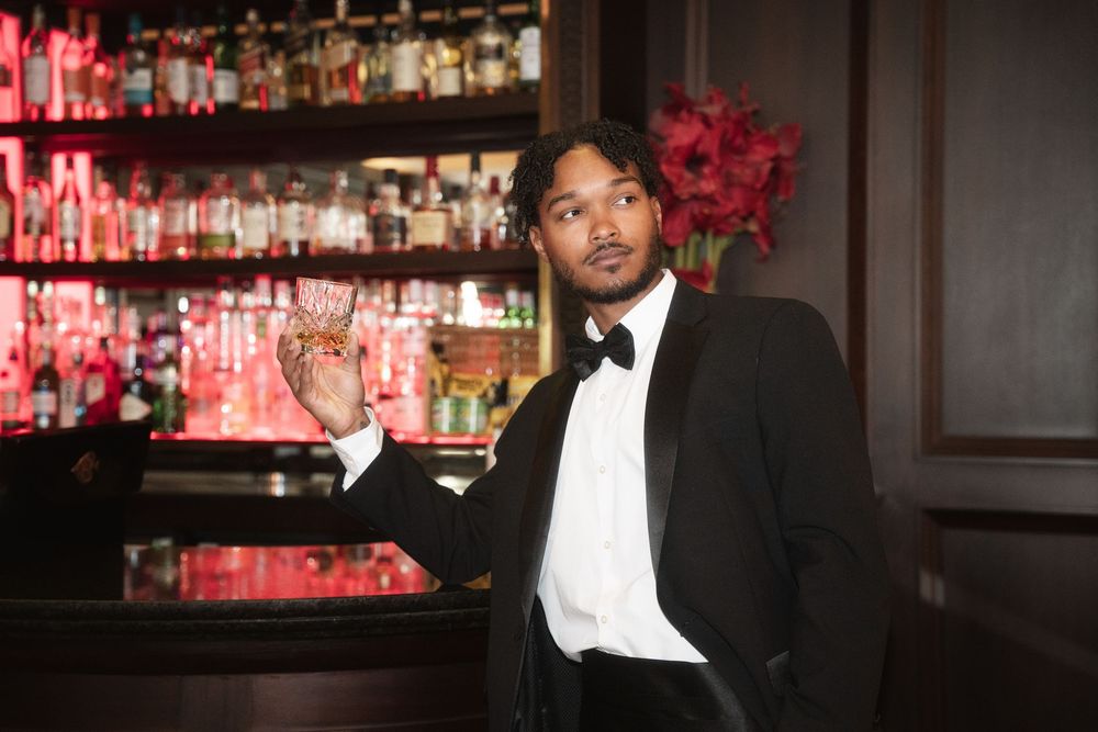 Elegant person in tuxedo standing at upscale bar with red lighting and bottles displayed behind them.