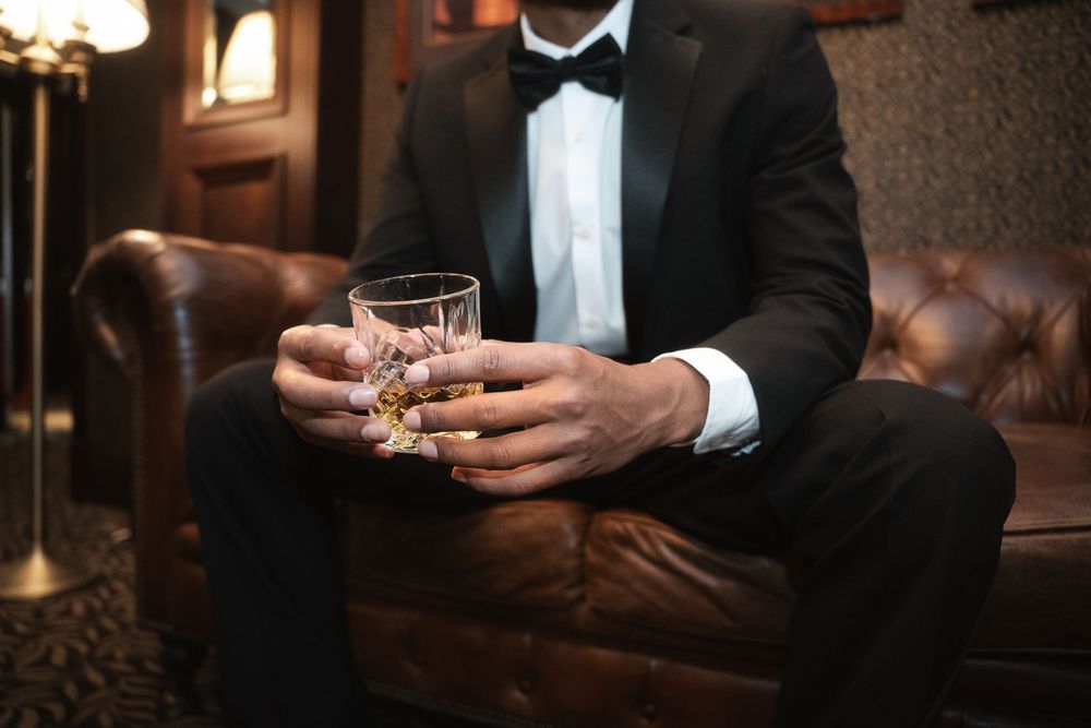 Person in formal black tuxedo sitting on leather chair holding crystal whiskey glass at upscale bar.