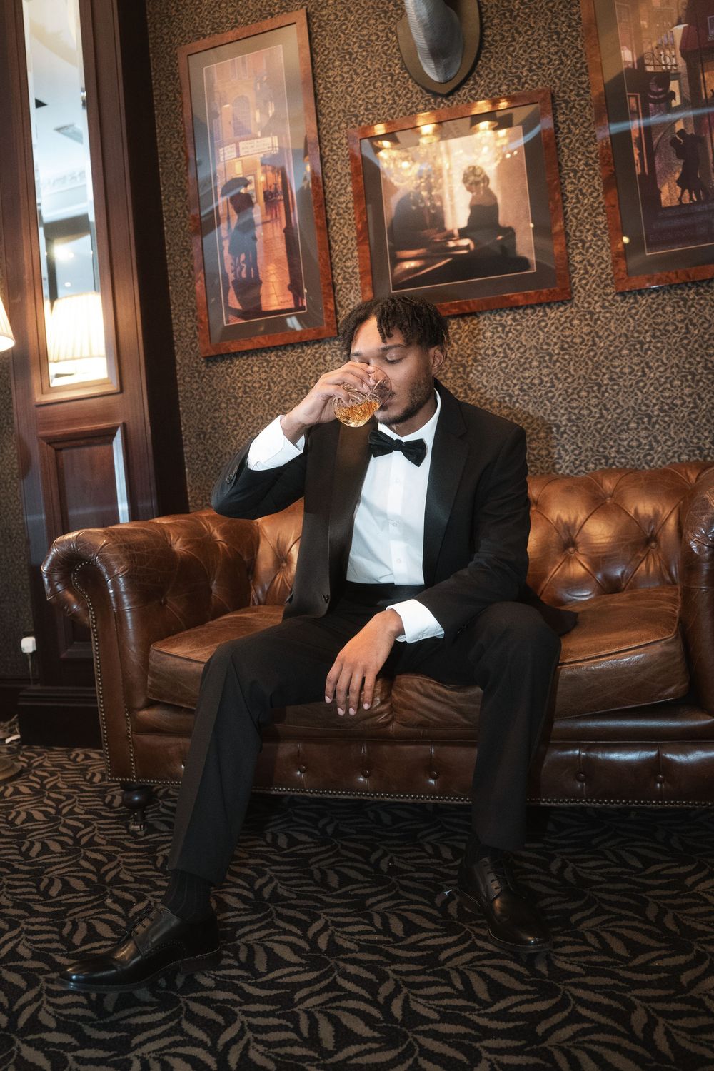 A person in formal attire enjoying a drink while seated on a vintage brown leather Chesterfield sofa in a stylish room.