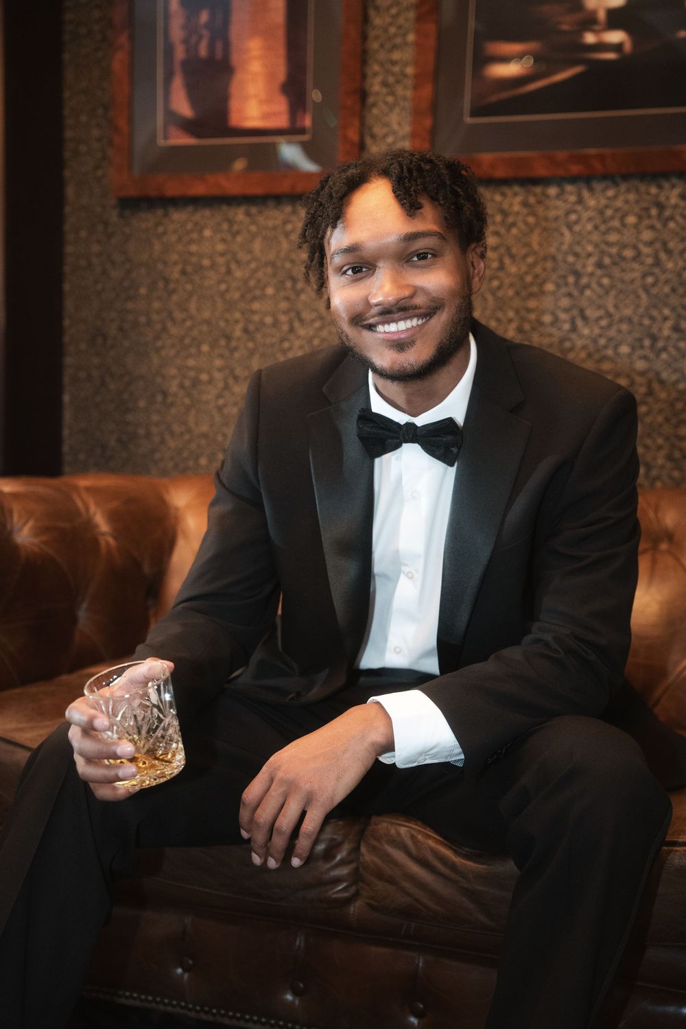 A well-dressed person in a black tuxedo and bowtie sits on a leather couch while holding a glass of whiskey.