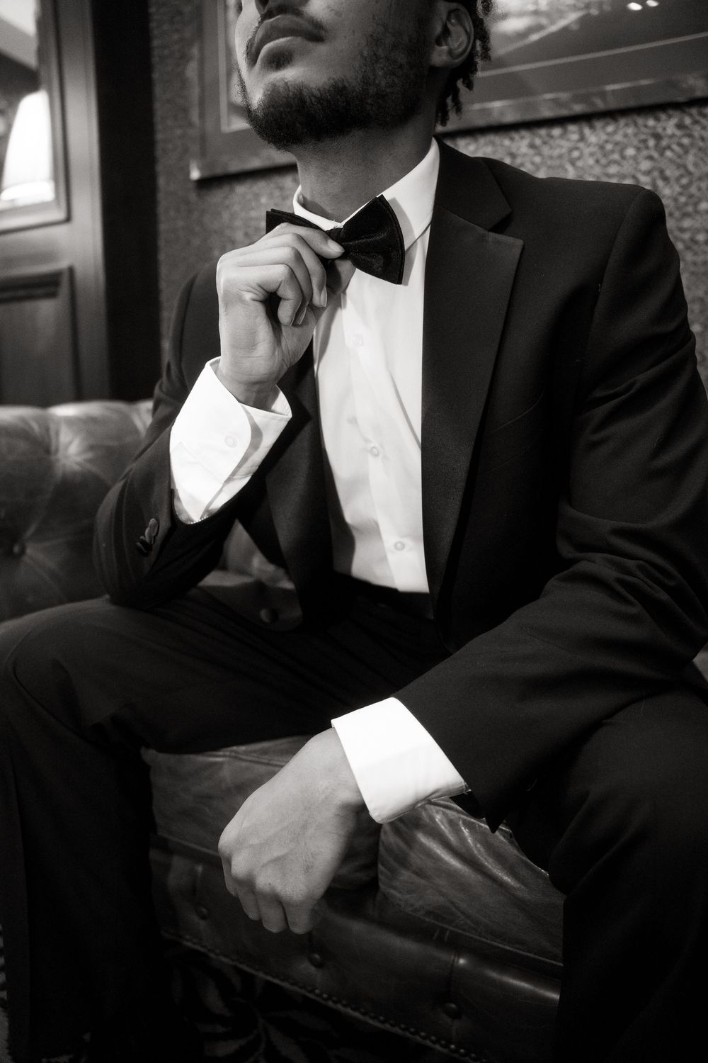 Black and white photo of person in tuxedo with bow tie sitting thoughtfully on leather chair in elegant setting.