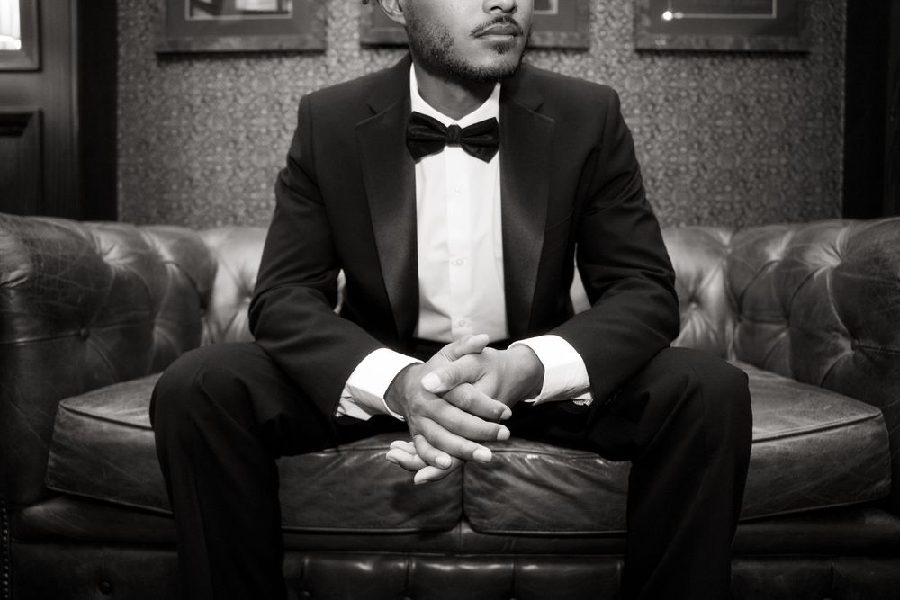 Person in formal black tuxedo sitting on dark leather couch while looking at smartphone screen.