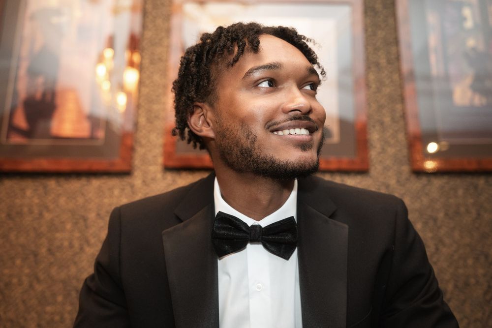 A person wearing a black tuxedo and bowtie smiles warmly in an elegant indoor setting.