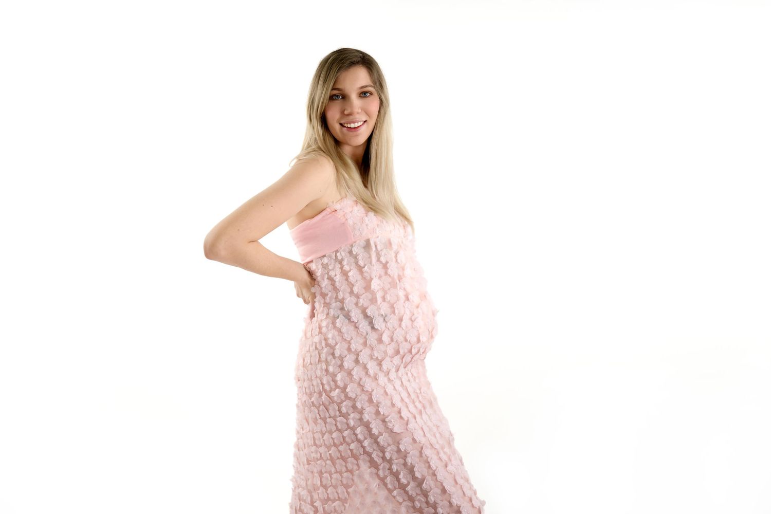A pregnant woman wearing a strapless pink textured dress poses against a white background.
