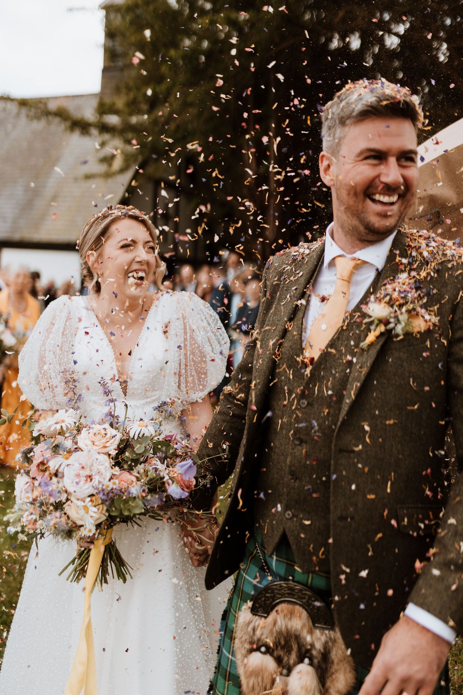 Joyful newlyweds laugh as confetti falls around them at outdoor wedding, bride holding purple bouquet and groom in kilt at Quaint Country