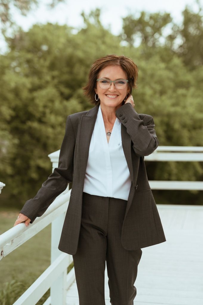 Professional dressed in dark business suit and white blouse poses outdoors on white bridge with a warm smile.