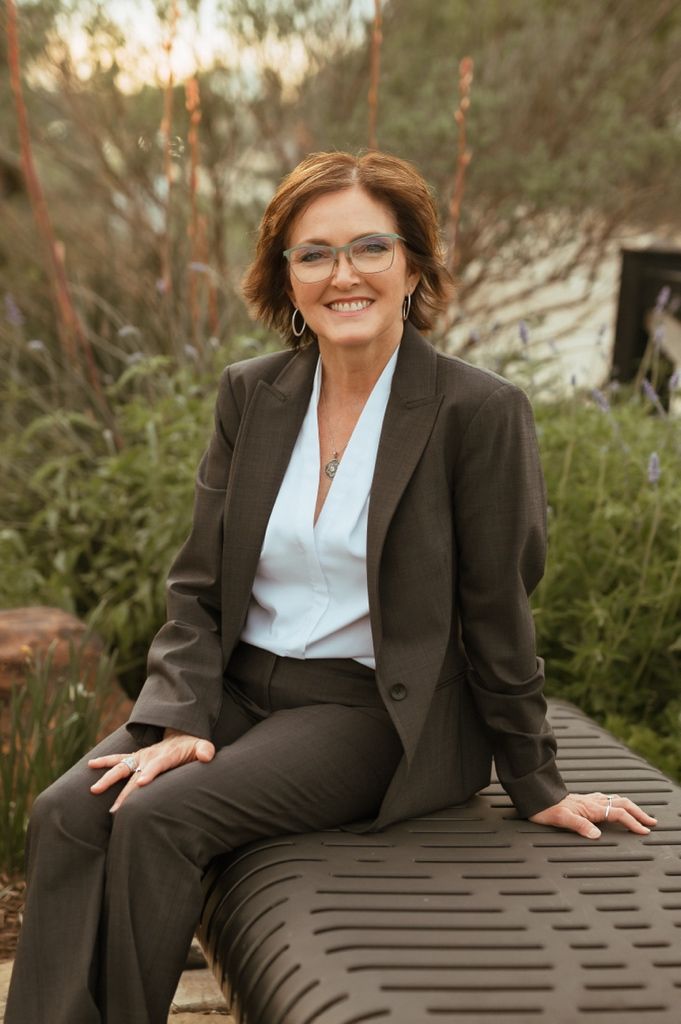 Professional portrait of someone in a brown business suit and glasses sitting outdoors at sunset with natural scenery behind.