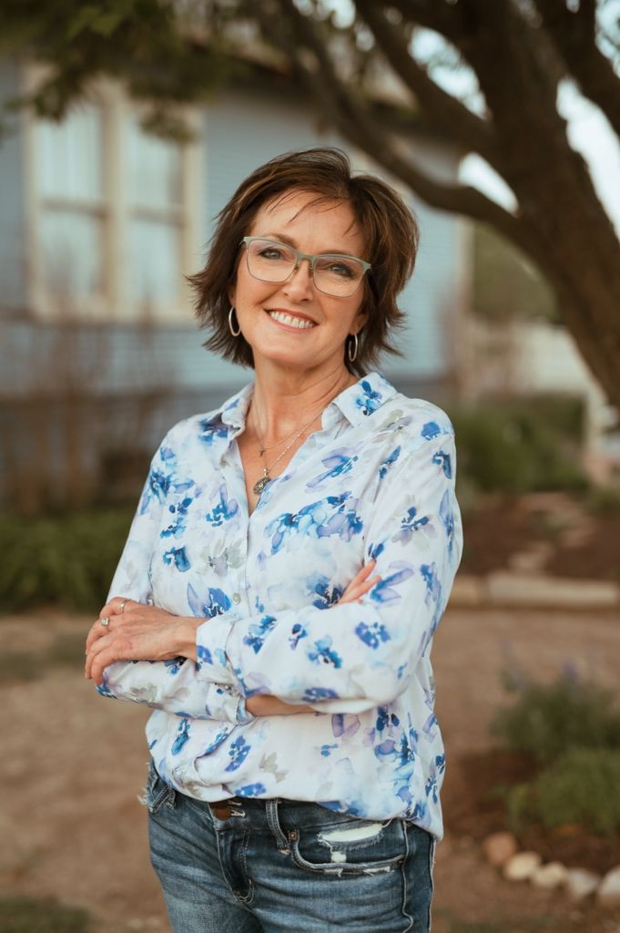 A warm portrait of someone wearing glasses and a blue floral print blouse with arms crossed, standing against a tree background.