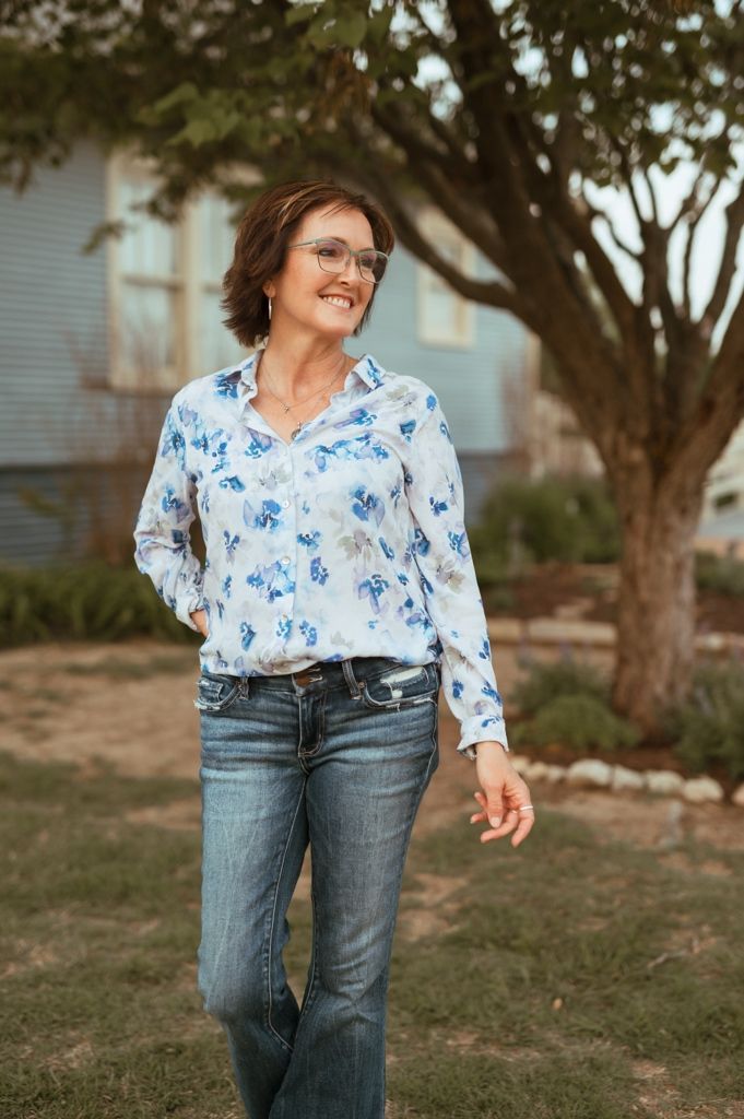 A person wearing glasses and a blue floral blouse smiles while standing outside near a tree.