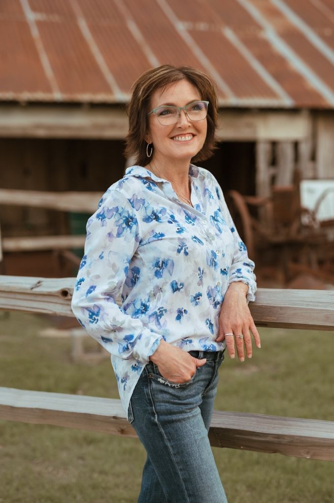 A woman wearing glasses and a blue floral blouse leans against a wooden fence near a rustic barn.