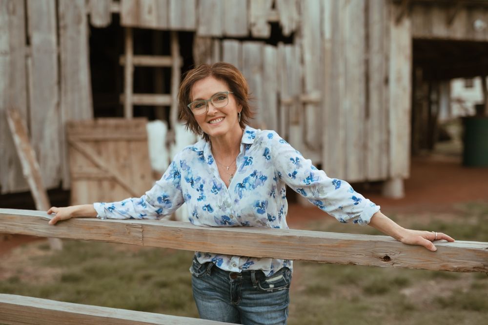 Someone wearing glasses and a blue floral shirt leans on a wooden fence rail near a rustic barn structure.