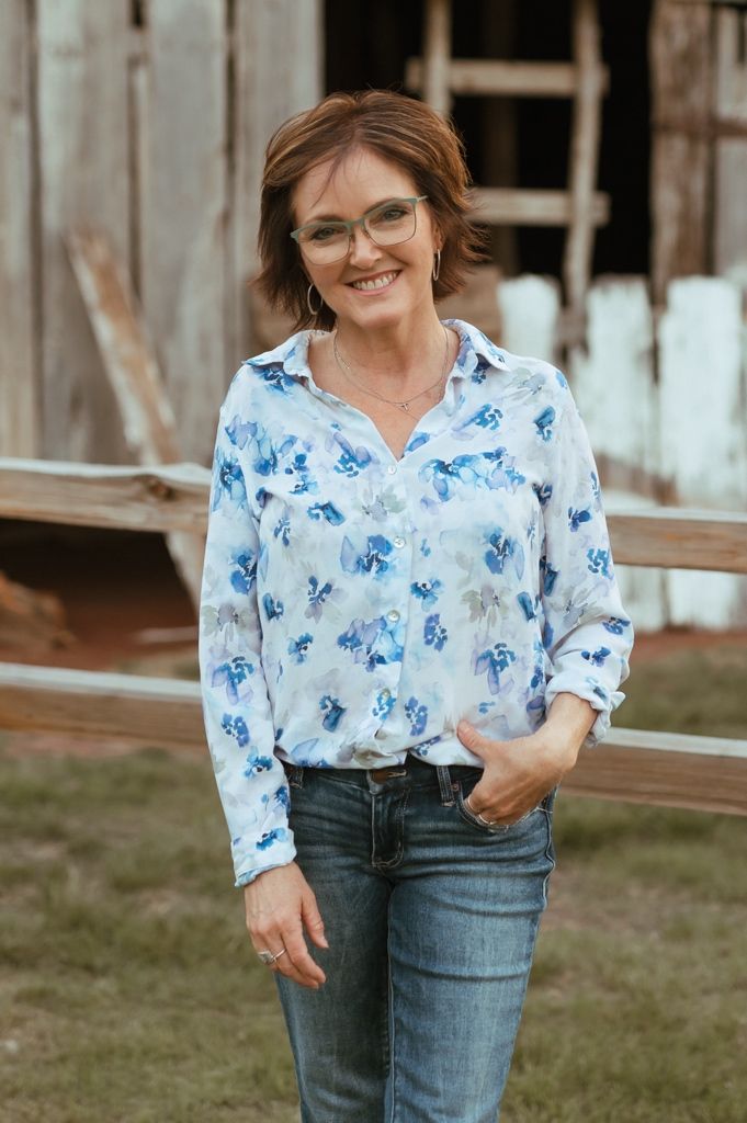 Woman wearing floral print blue and white top with glasses and jeans poses casually by rustic wooden fence.