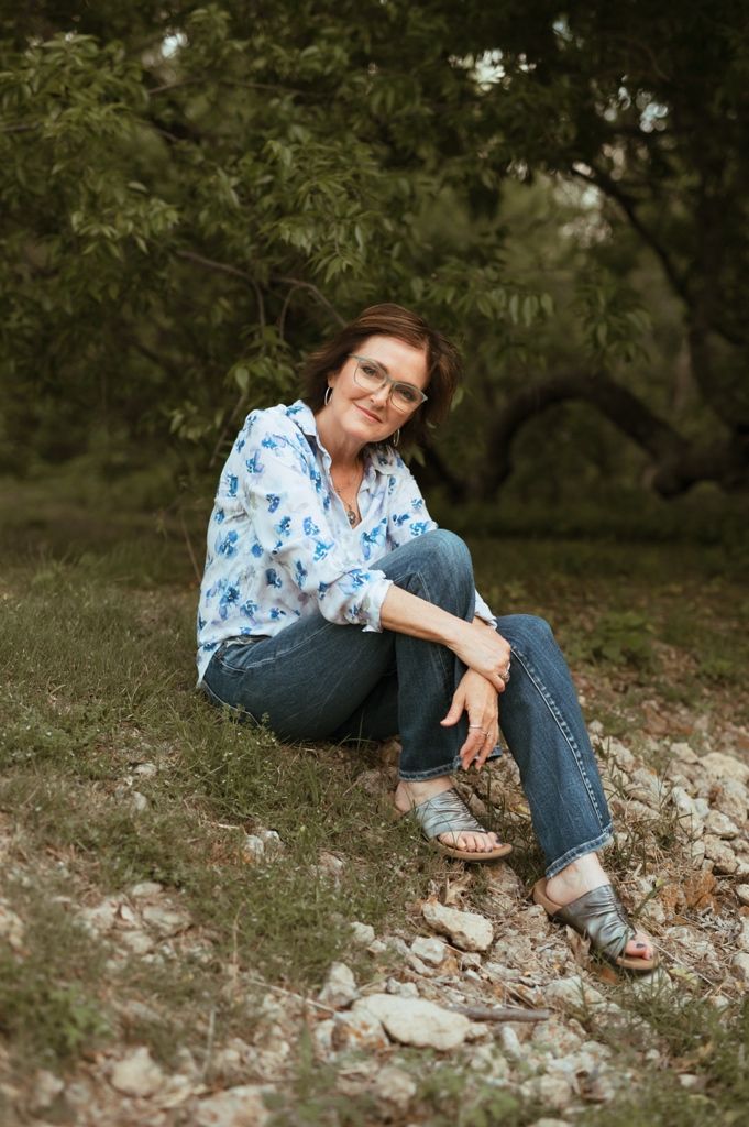 Someone relaxes outdoors on rocky ground wearing a floral blouse and jeans against a leafy green backdrop.